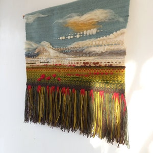 Puede incluir: Tapiz de pared tejido a mano que representa un paisaje. Presenta un cielo azul con nubes blancas y amarillas, un paisaje texturizado en tonos verdes, rojos y marrones, y una franja inferior con hilos amarillos, verdes y rojos. Montado en una barra de madera.