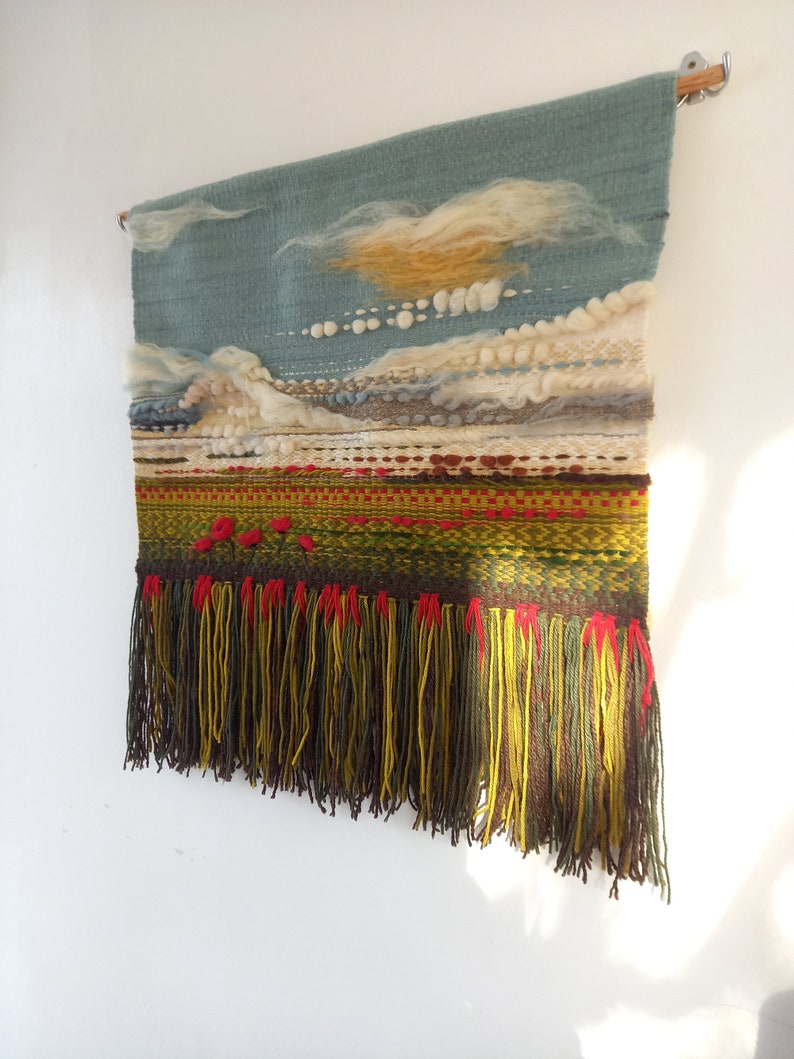 Puede incluir: Tapiz de pared tejido a mano que representa un paisaje. Presenta un cielo azul con nubes blancas y amarillas, un paisaje texturizado en tonos verdes, rojos y marrones, y una franja inferior con hilos amarillos, verdes y rojos. Montado en una barra de madera.
