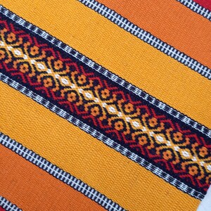 May include: A close-up of a woven fabric with a repeating pattern of orange, red, black, and white stripes. The fabric has a textured surface and a geometric design.