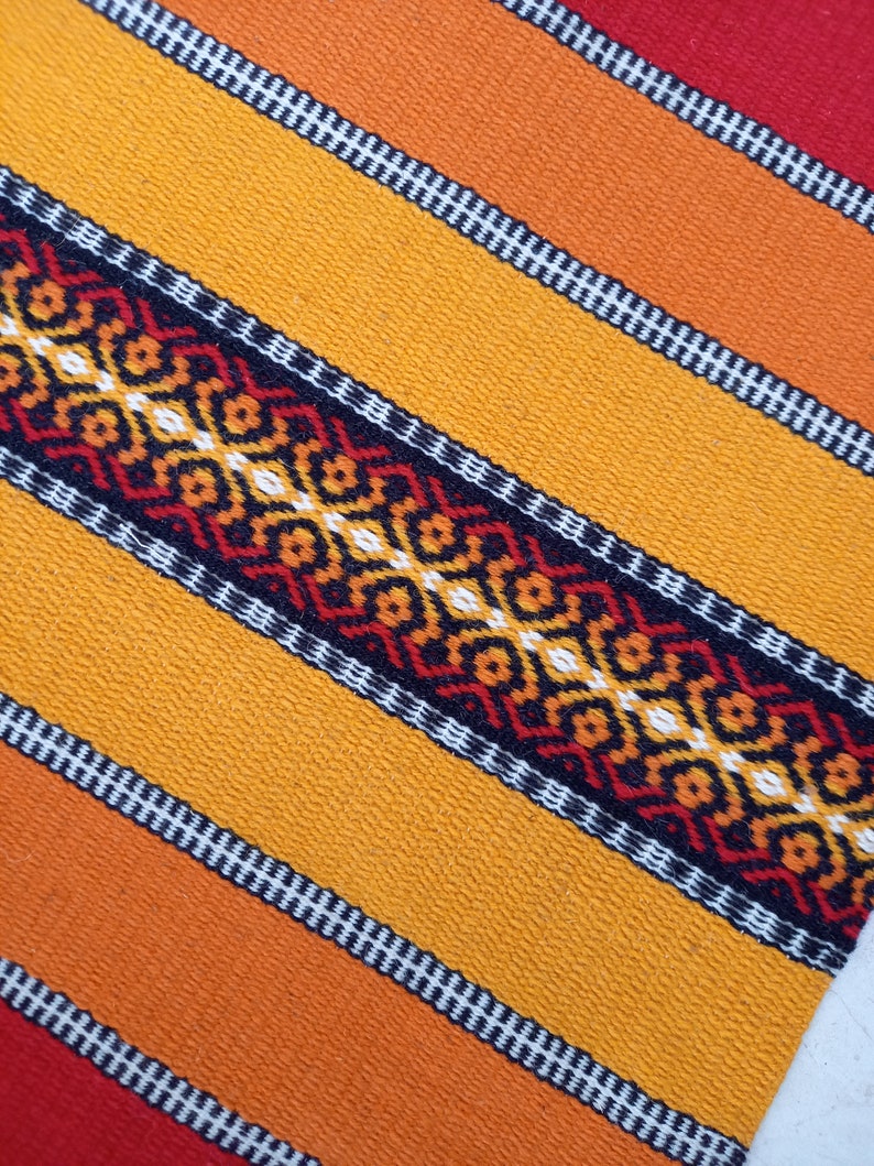 May include: A close-up of a woven fabric with a repeating pattern of orange, red, black, and white stripes. The fabric has a textured surface and a geometric design.