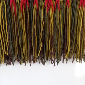 Puede incluir: Primer plano de un textil tejido con un borde con flecos. La parte superior presenta un patr&oacute;n geom&eacute;trico en tonos marrones, verdes y rojos. La franja consta de largos hilos en verde oliva, marr&oacute;n y amarillo, con detalles en rojo.