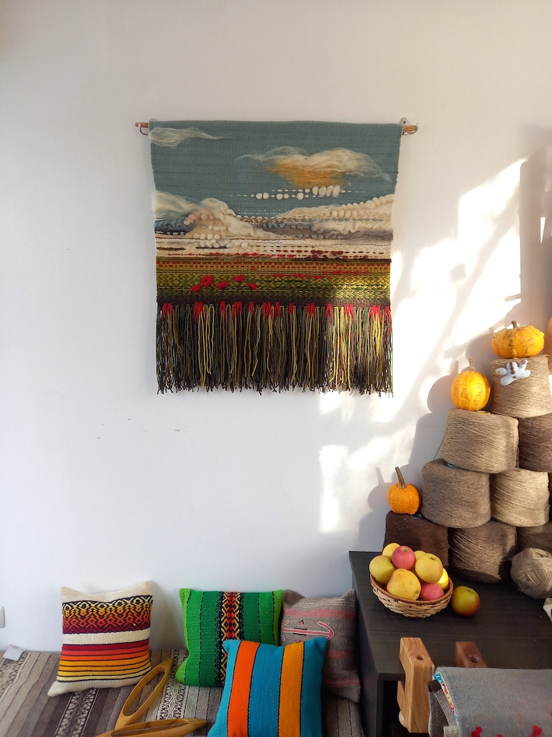 Puede incluir: Un tapiz tejido que representa un paisaje con cielo azul, nubes y un campo de flores rojas. El tapiz tiene una franja en la parte inferior. Cojines coloridos y una cesta de manzanas en primer plano.