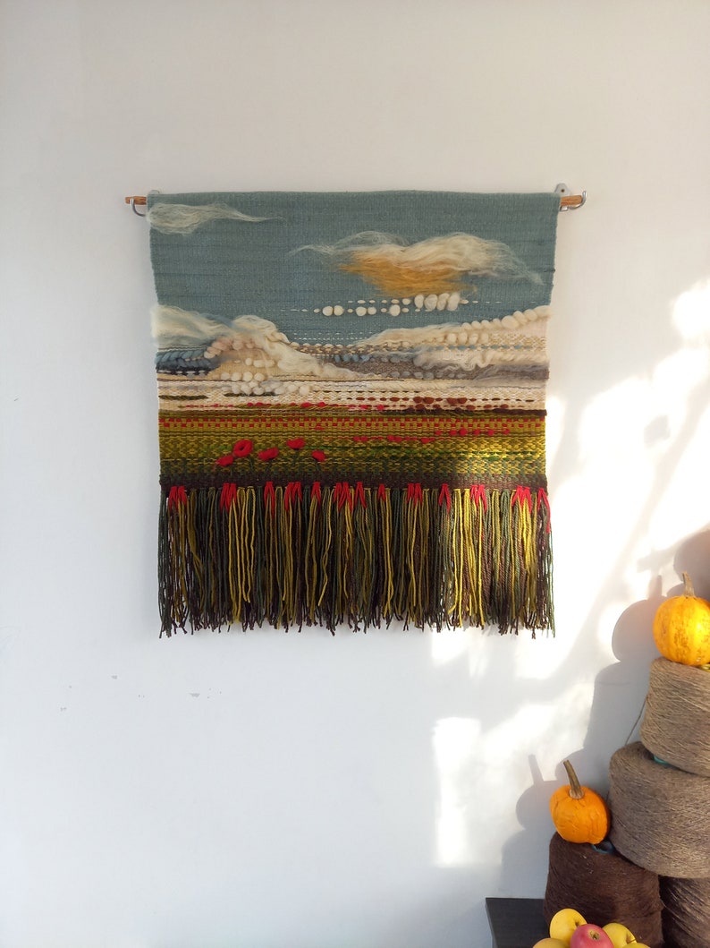 Puede incluir: Tapiz de pared tejido a mano que representa un paisaje con cielo azul, nubes blancas y amarillas, y un campo verde y rojo. El borde inferior presenta largas borlas con flecos en tonos marr&oacute;n, amarillo y rojo. El tapiz est&aacute; montado en una barra de madera.