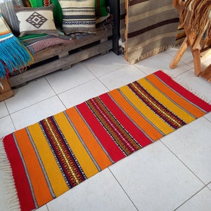 May include: A colorful handwoven rug with red, orange, and yellow stripes and black geometric patterns. The rug has white fringe on the ends.