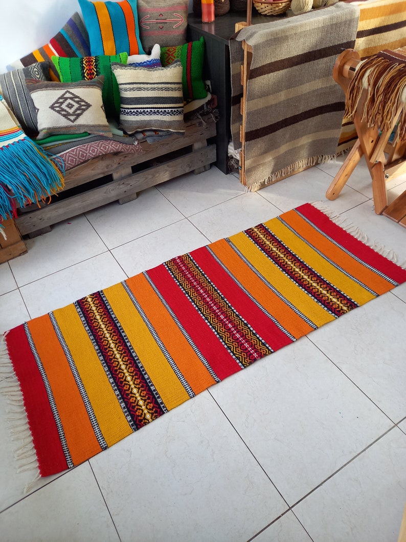 May include: A colorful handwoven rug with red, orange, and yellow stripes and black geometric patterns. The rug has white fringe on the ends.