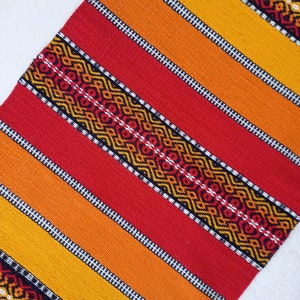 May include: A woven rug with a geometric pattern in shades of red, orange, and yellow. The rug has a black and white border.