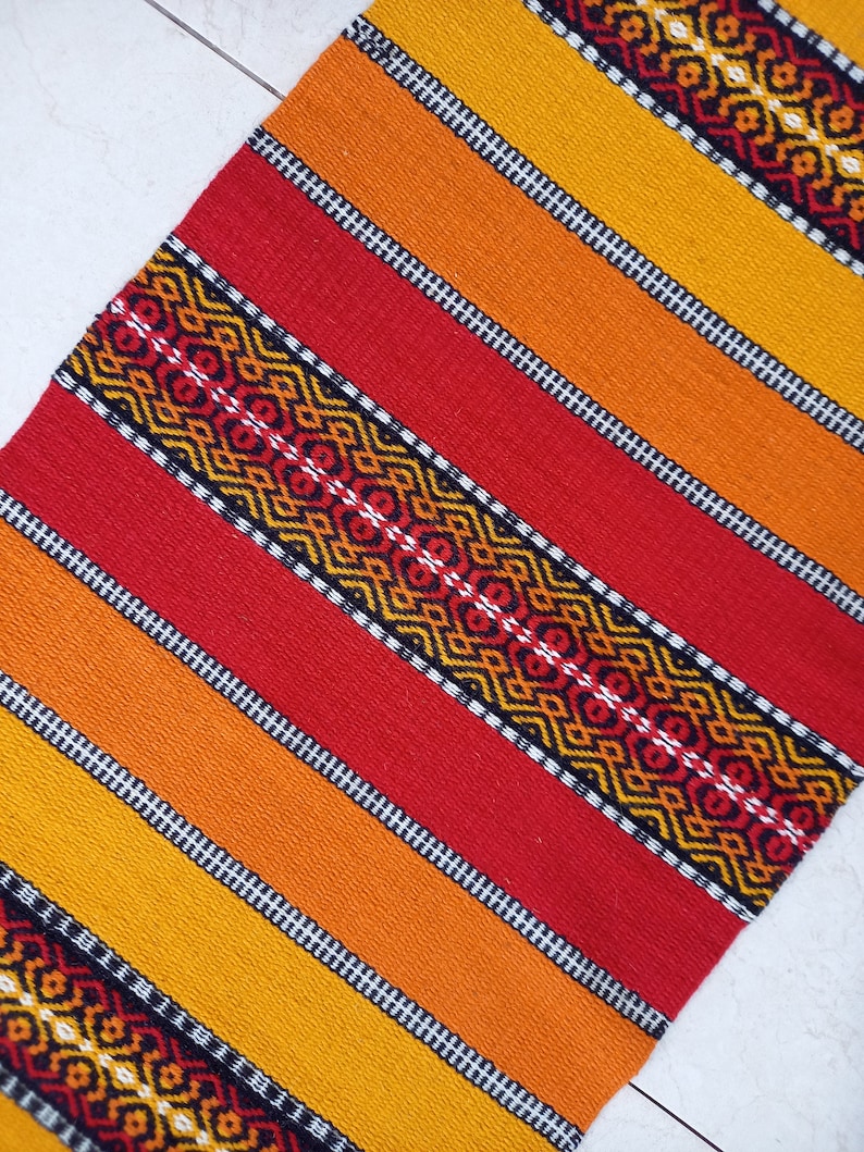 May include: A woven rug with a geometric pattern in shades of red, orange, and yellow. The rug has a black and white border.