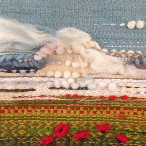 Puede incluir: Una obra de arte textil tejida que representa una escena de paisaje. La parte superior presenta un cielo azul claro con nubes blancas y esponjosas. Debajo, un paisaje texturizado con elementos verdes, rojos y marrones, incluyendo acentos de flores rojas. La parte inferior tiene una franja.