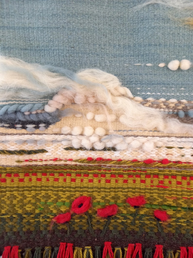 Puede incluir: Una obra de arte textil tejida que representa una escena de paisaje. La parte superior presenta un cielo azul claro con nubes blancas y esponjosas. Debajo, un paisaje texturizado con elementos verdes, rojos y marrones, incluyendo acentos de flores rojas. La parte inferior tiene una franja.