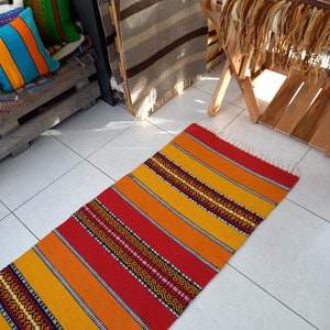 May include: A long, narrow, handwoven rug with alternating stripes of red, orange, and yellow. The rug has a geometric pattern woven into the stripes.