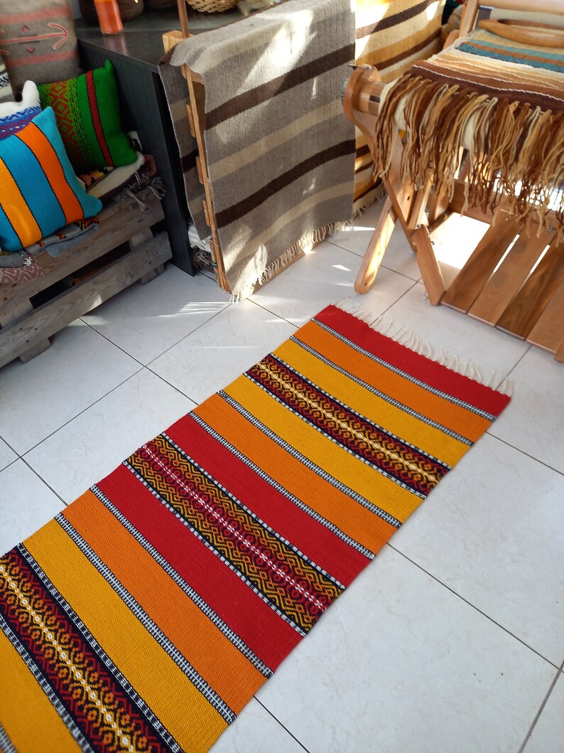 May include: A long, narrow, handwoven rug with alternating stripes of red, orange, and yellow. The rug has a geometric pattern woven into the stripes.