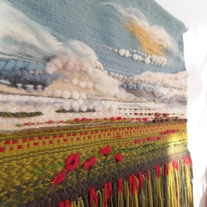 Puede incluir: Tapiz de pared tejido a mano con un dise&ntilde;o de paisaje. La parte superior presenta un cielo azul con nubes blancas y amarillas. La parte inferior tiene un campo verde con detalles de flores rojas y una franja de hilos amarillos y rojos.