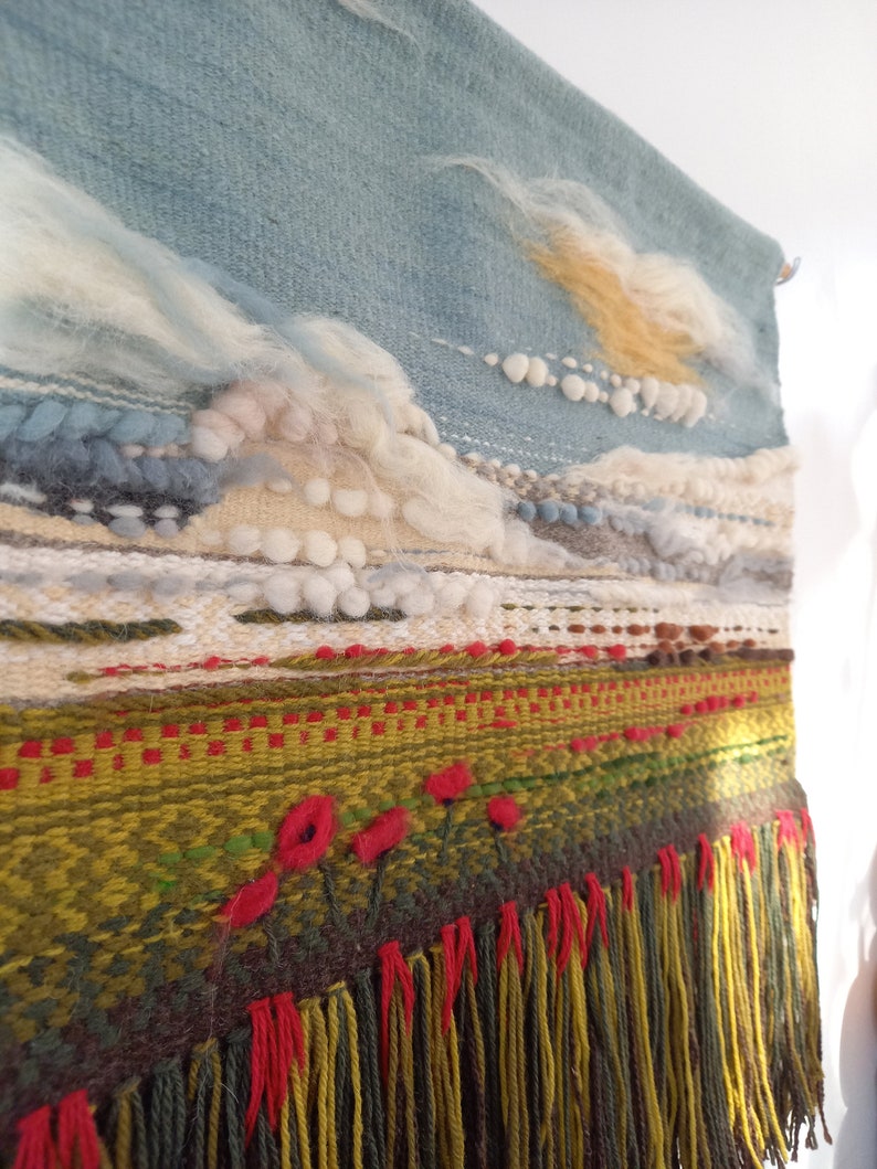 Puede incluir: Tapiz de pared tejido a mano con un dise&ntilde;o de paisaje. La parte superior presenta un cielo azul con nubes blancas y amarillas. La parte inferior tiene un campo verde con detalles de flores rojas y una franja de hilos amarillos y rojos.