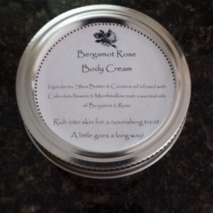 May include: A silver lid with a white label that reads "Bergamot Rose Body Cream". The label lists the ingredients: Shea Butter & Coconut oil infused with Calendula flowers & Marshmallow root, essential oils of Bergamot & Rose. The label also says "Rub into skin for a nourishing treat. A little goes a long way!"