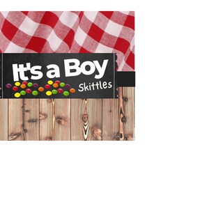 May include: A chalkboard sign with the text "It's a Boy" and the Skittles logo. The sign is decorated with colorful Skittles candies and is set against a rustic wooden background.
