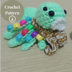 Puede incluir: Un pulpo verde de crochet con ojos negros y una bufanda blanca de crochet. El pulpo tiene cuentas de colores en sus tentáculos.