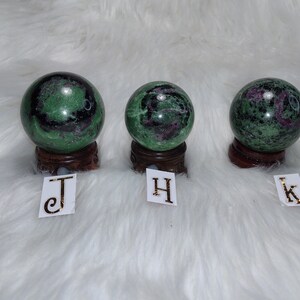May include: Three green, black, and pink gemstone spheres on wooden stands. The spheres are labelled J, H, and K.