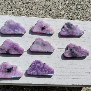 May include: Eight purple and black cloud-shaped stones on a white wooden surface. Each stone has a small white label with a number on it.
