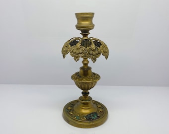 May include: A gold-toned metal candlestick with an ornate design. The base is round and has a decorative pattern. The candlestick has a central pillar with a cup for holding a candle.