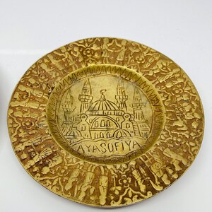 May include: A gold-colored metal plate with an intricate design. The center of the plate features a depiction of the Hagia Sophia mosque in Istanbul, Turkey. The text "AYASOFYA" is inscribed below the mosque.