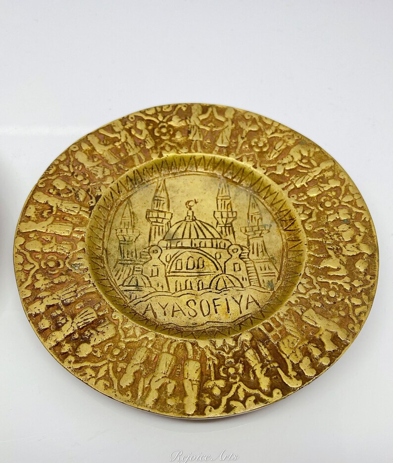 May include: A gold-colored metal plate with an intricate design. The center of the plate features a depiction of the Hagia Sophia mosque in Istanbul, Turkey. The text "AYASOFYA" is inscribed below the mosque.