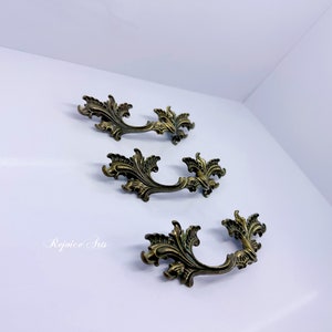May include: Three ornate, gold-toned metal drawer pulls with a leaf design. The pulls have a curved shape and are designed to be used on cabinets or furniture.