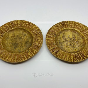 May include: Two small, round, gold-colored metal plates with an intricate design around the edges. The center of each plate features an engraved image of the Hagia Sophia mosque in Istanbul, Turkey. The text "AYA SOFIYA" is engraved on one of the plates.