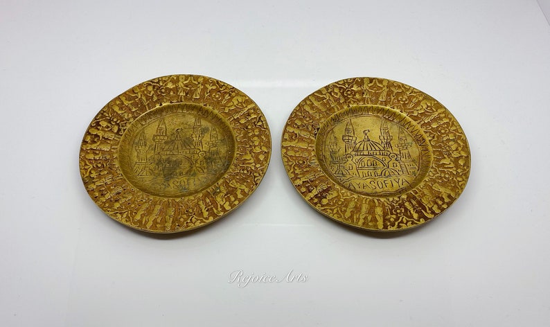 May include: Two small, round, gold-colored metal plates with an intricate design around the edges. The center of each plate features an engraved image of the Hagia Sophia mosque in Istanbul, Turkey. The text "AYA SOFIYA" is engraved on one of the plates.