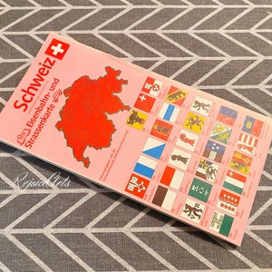 May include: A pink and white map of Switzerland with a red outline of the country. The map is titled "Schweiz Eisenbahn- und Strassenkarte" and features a small Swiss flag in the top right corner. The map also includes a key with small flags representing different cantons.