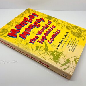 Puede incluir: Un libro amarillo titulado "Manga! El mundo del c&oacute;mic japon&eacute;s" de Frederik L. Schodt. La portada presenta ilustraciones de personajes de manga y el texto "Pr&oacute;logo de Osamu Tezuka".