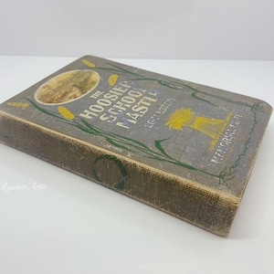 May include: A vintage book titled "The Hoosier School Master" by Edward Eggleston. The cover features a blue background with illustrations of wheat stalks, a landscape, and a farmhouse.