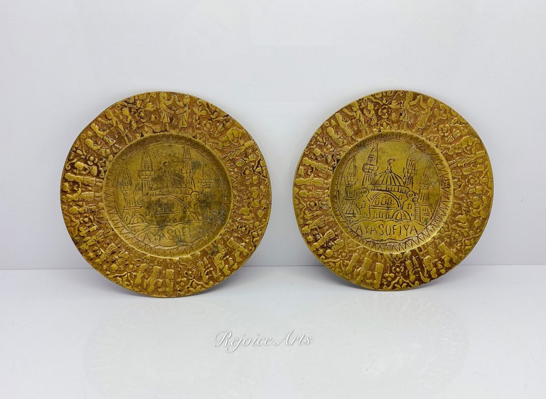 May include: Two small, round, gold-colored brass plates with an embossed design. The plates feature a depiction of the Hagia Sophia mosque in Istanbul, Turkey. The text "AYA SOFİYA" is inscribed on the plates.