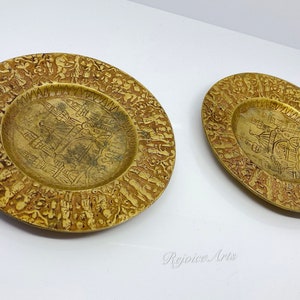 May include: Two gold-colored metal plates with intricate designs. The plates have a raised rim and a center design of a building.
