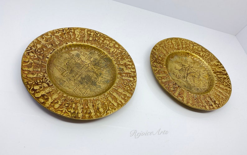 May include: Two gold-colored metal plates with intricate designs. The plates have a raised rim and a center design of a building.