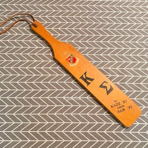 May include: A wooden paddle with a leather strap, featuring the letters "KΣ" and the text "TO KAZZ '57 FROM SAM '57".
