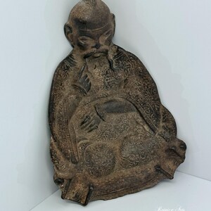 May include: A dark brown, textured metal wall plaque featuring a relief sculpture of a seated figure with a mustache and traditional headwear. The figure wears ornate, patterned robes and holds a small object. The text Rejoice Arts is visible.