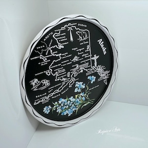May include: A circular, black tray with a white, scalloped border, displaying a white map of Alaska. The tray includes illustrations of a bear, mountains, and flowers. The word "Alaska" is written in white on the right side.