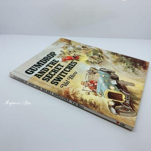 May include: A hardcover book titled "Gumdrop and the Secret Switches" by Val Biro. The cover features an illustration of a vintage car and other vehicles. The book is set against a white background.