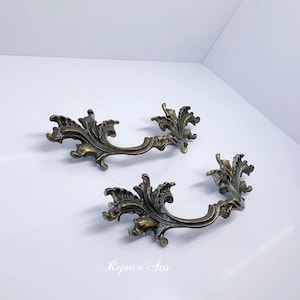 May include: Two ornate brass drawer pulls with a floral design. The pulls are in the shape of a half-circle with a decorative scrollwork design.