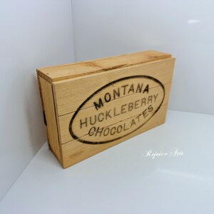 May include: A wooden box with a lid, featuring the text "MONTANA HUCKLEBERRY CHOCOLATES" in a black oval design. The box is made of light-colored wood and has a rustic, vintage aesthetic. The box is rectangular and appears to be empty.