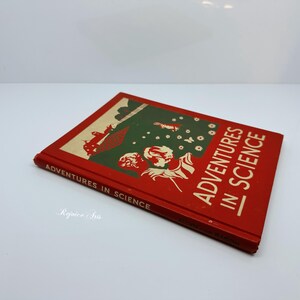 May include: A vintage book titled "Adventures in Science" with a red cover. The cover features illustrations in green and white, with the title in large white letters. The book is angled on a white surface.