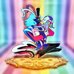 May include: A colorful acrylic stand featuring two cartoon characters embracing. One character has blue hair and a top hat, the other is wearing a jester-like outfit. The stand has a gold-colored base with an ornate design and a rainbow background.