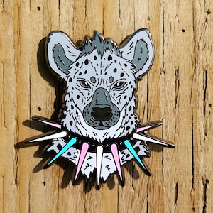 May include: An enamel pin depicting a hyena's head, adorned with a spiked collar. The hyena is grey with black spots and a black nose. The collar features pink and blue spikes. The pin is set against a wooden background.