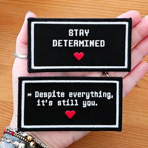 May include: Two black and white embroidered patches with red hearts. The top patch says "Stay Determined" and the bottom patch says "* Despite everything, it's still you."