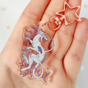 Op de afbeelding: Een transparante sleutelhanger met een wit, iriserend wezen met een lange staart, omringd door roze en paarse bloemdetails. De sleutelhanger is bevestigd aan een roségouden stervormige hanger met een metalen ring.