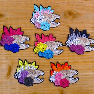 Puede incluir: Seis parches bordados con un diseño de calavera adornados con detalles de cristales y flores de colores. Cada parche presenta una combinación de colores diferente, incluyendo rosa, azul, amarillo y naranja, sobre una calavera marrón.