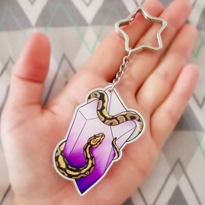 Może przedstawiać: Zawieszka z fioletowego kryształu z owiniętym wokół niej żółto-brązowym wężem. Zawieszka jest przymocowana do srebrnego breloczka w kształcie gwiazdy.