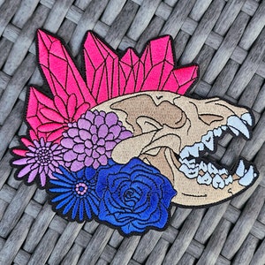 May include: An embroidered patch featuring a stylised animal skull with pink, purple and blue flowers and crystals. The skull is beige with white teeth and the flowers are in shades of pink, purple and blue.