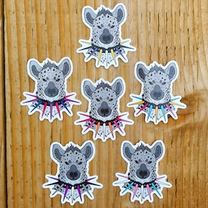 May include: Six stickers of hyena heads, each with a studded collar. The hyenas are grey with black spots. The collars feature spikes in pink, blue, yellow, and purple. The stickers are on a wooden background.
