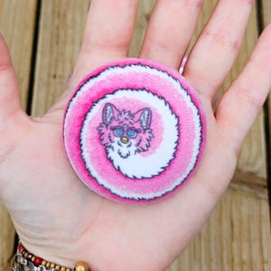 May include: A round, pink and white pin with a spiral design and a cartoon Furby face in the center. The pin is held in a hand, and the background is a wooden surface.